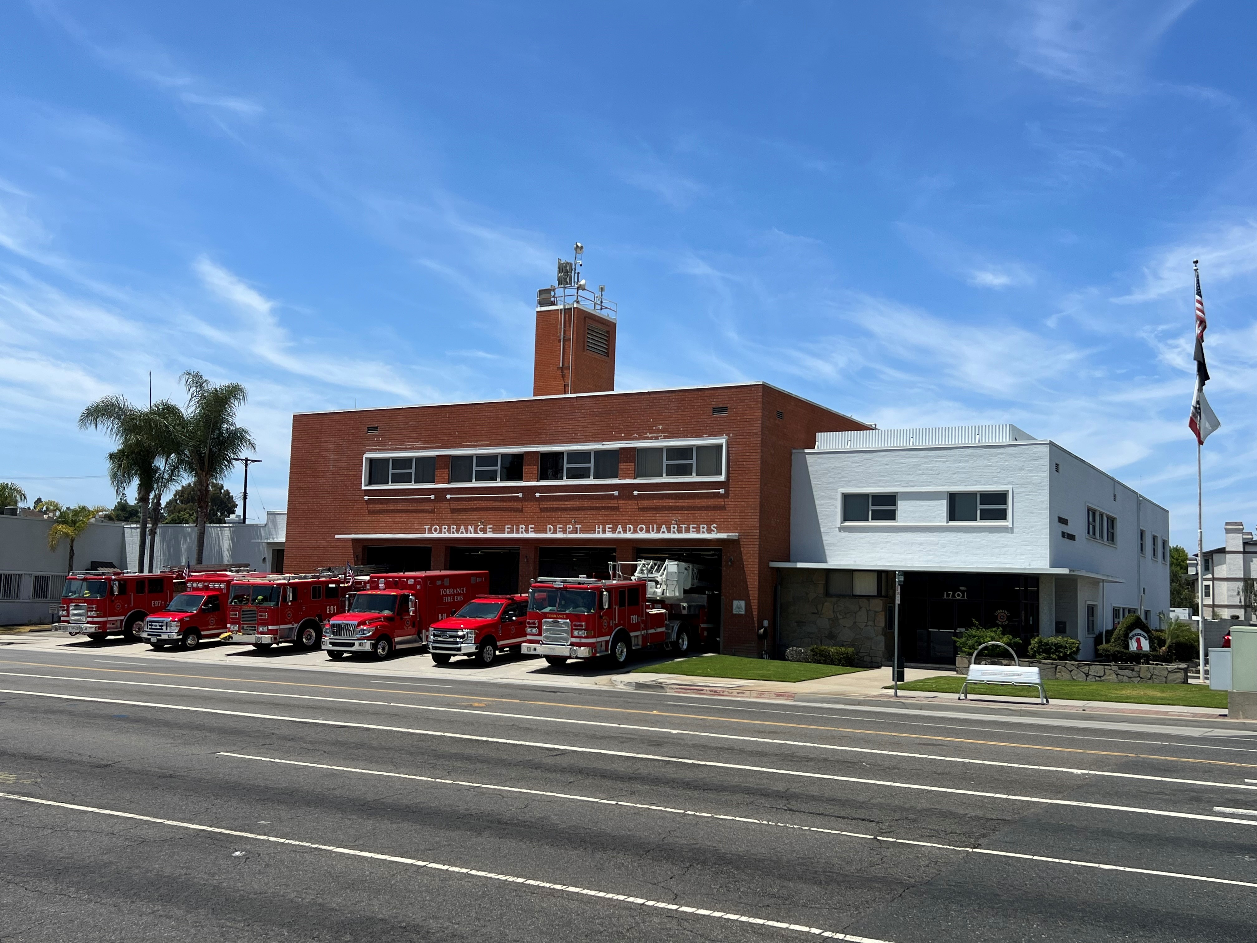A row of red fire trucks parked in front of a brick fire station building along a sunny street.