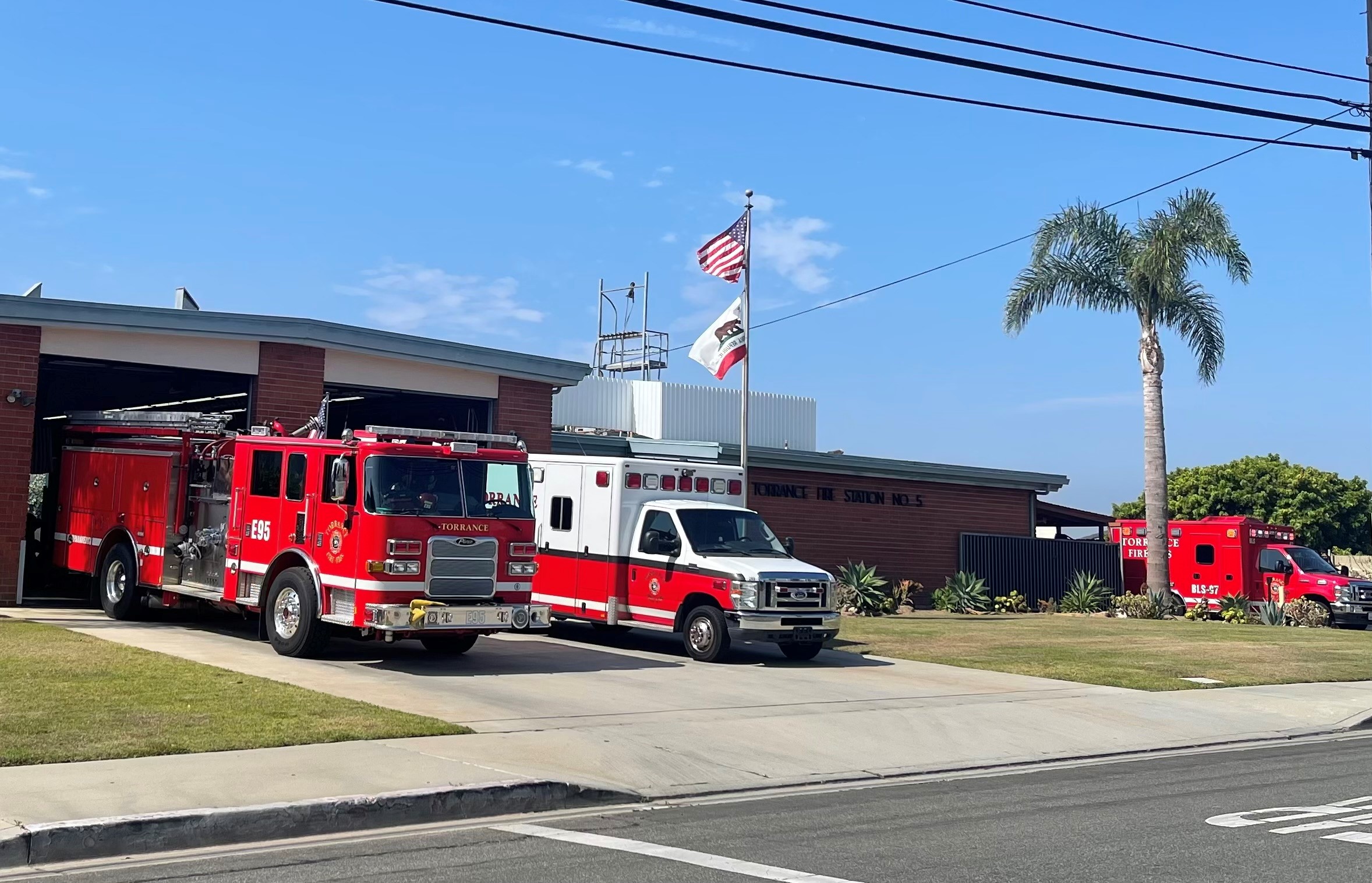 Daytime view of a fire station with a fire engine and ambulance parked outside, American and California flags flying above, with a palm tree nearby under a clear blue sky.