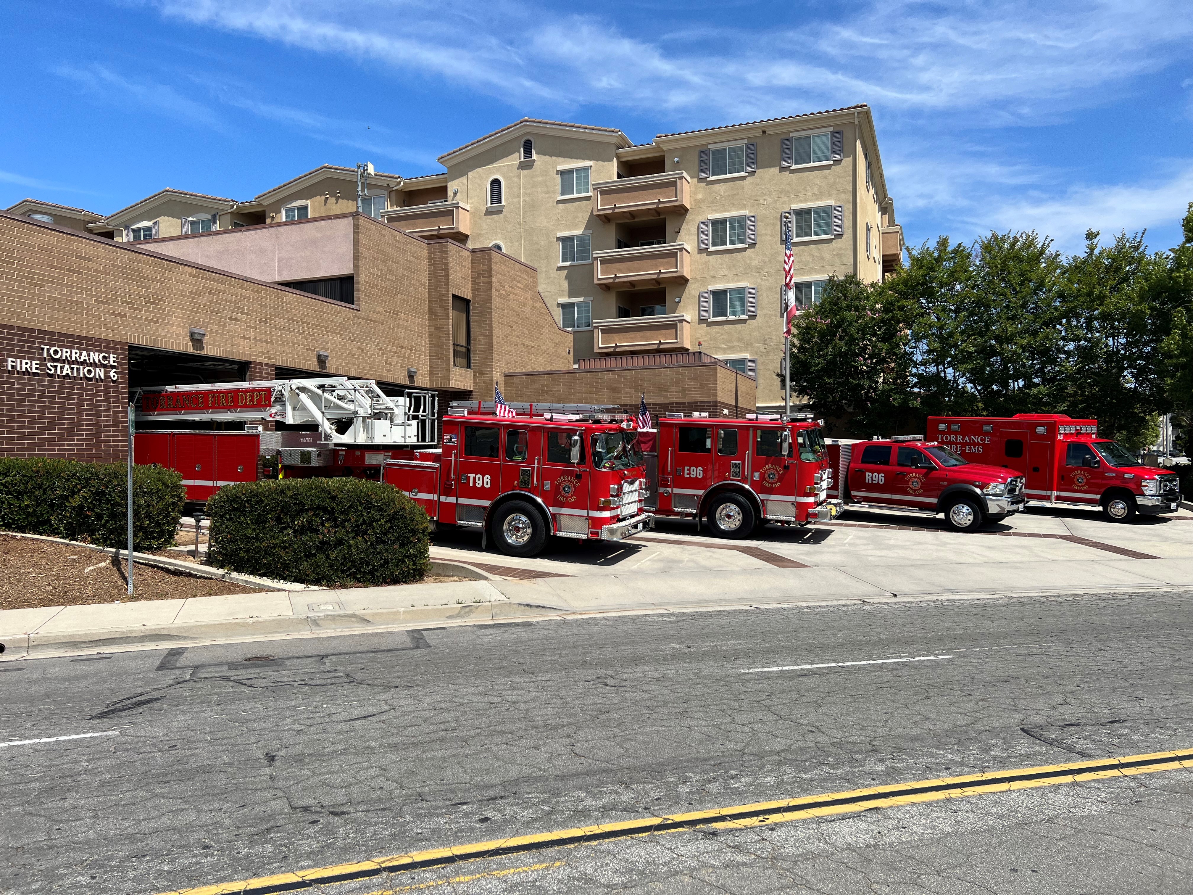 **Suggested alt text:** Fire station with multiple red fire engines, including a ladder truck, parked outside along the street in front of a multi-story residential building under a blue sky.