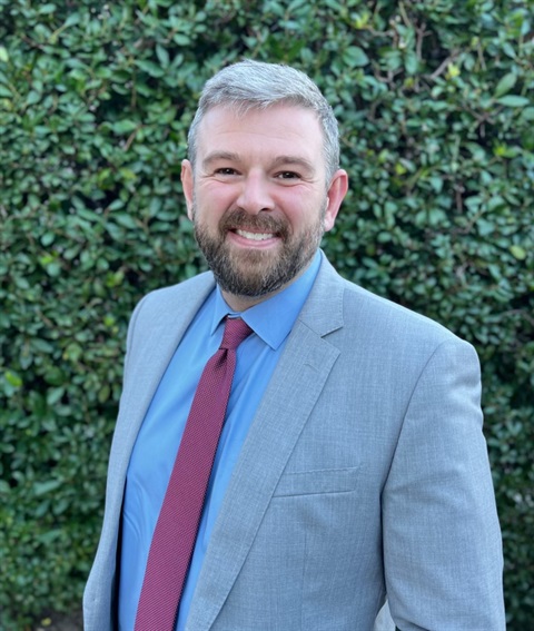 Professional headshot of Ian Dailey smiling in front of a leafy green background.